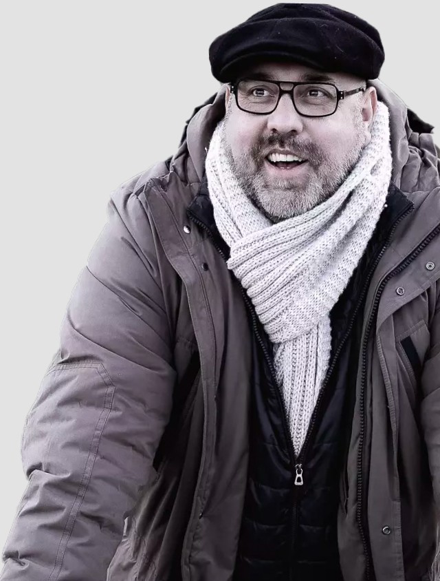 Ein Bild mit grauem Hintergrund und Roland Wagenhäuser von vorne. Er strahlt und richtet seinen Blick nach vorne. Er trägt die braune Jacke, den dicken weißen Wollschal und seine dunkle Brille und Baskenmütze. Roland nimmt fast das ganze Bild ein.