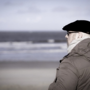 Das Bild zeigt Roland seitlich von hinten mit Blick auf das Meer. Die Wellen sind verschwommen und Rolands Blick geht leicht nach links, so dass man sein Profil mit schwarzer Baskenmütze, Brille, linkem Ohr und der Kapuze von der Jacke sitz. Die Jacke ist braun und wirkt sehr gemütlich.