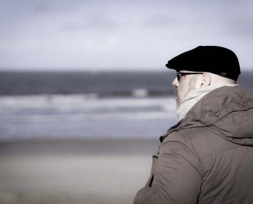 Das Bild zeigt Roland seitlich von hinten mit Blick auf das Meer. Die Wellen sind verschwommen und Rolands Blick geht leicht nach links, so dass man sein Profil mit schwarzer Baskenmütze, Brille, linkem Ohr und der Kapuze von der Jacke sitz. Die Jacke ist braun und wirkt sehr gemütlich.