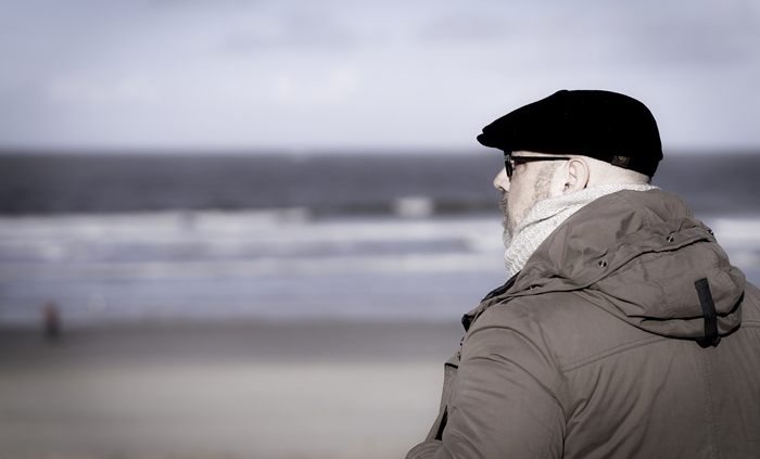 Das Bild zeigt Roland seitlich von hinten mit Blick auf das Meer. Die Wellen sind verschwommen und Rolands Blick geht leicht nach links, so dass man sein Profil mit schwarzer Baskenmütze, Brille, linkem Ohr und der Kapuze von der Jacke sitz. Die Jacke ist braun und wirkt sehr gemütlich.