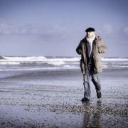 Roland Wagenhäuser geht im Wasserraum entlang. Hinten sieht man die Wellen an den Strand kommen, und Roland geht über Muscheln und Steine über den Strand. Der Himmel ist blau mit Wolen und Roland ist in Bewegung. Er trägt eine Jeans, die dicke braune Jacke und den weißen Wollschal. Man erahnt die Brille und sieht die schwarze Baskenmütze.