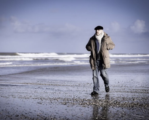 Roland Wagenhäuser geht im Wasserraum entlang. Hinten sieht man die Wellen an den Strand kommen, und Roland geht über Muscheln und Steine über den Strand. Der Himmel ist blau mit Wolen und Roland ist in Bewegung. Er trägt eine Jeans, die dicke braune Jacke und den weißen Wollschal. Man erahnt die Brille und sieht die schwarze Baskenmütze.
