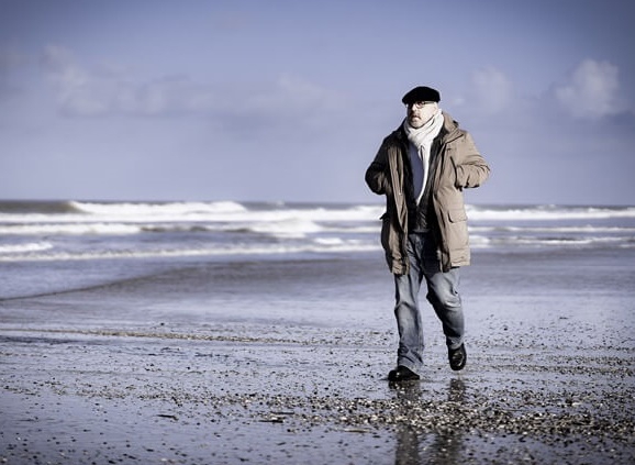 Roland Wagenhäuser geht im Wasserraum entlang. Hinten sieht man die Wellen an den Strand kommen, und Roland geht über Muscheln und Steine über den Strand. Der Himmel ist blau mit Wolen und Roland ist in Bewegung. Er trägt eine Jeans, die dicke braune Jacke und den weißen Wollschal. Man erahnt die Brille und sieht die schwarze Baskenmütze.