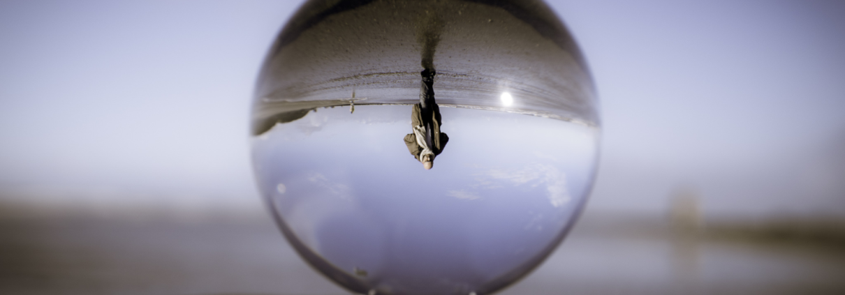Das Bild zeigt einen Lensball, der alle Bilder auf den Kopf stellt. Er ist in der Mitte des Bildes und steht auf einem Holzpoller. Bis auf das Bild in der Kugel ist alles verschwommen. Dort sieht man Roland über den Strand gehen mit einem Blick nach vorne. Er steht dabei Kopf.