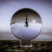 Das Bild zeigt einen Lensball, der alle Bilder auf den Kopf stellt. Er ist in der Mitte des Bildes und steht auf einem Holzpoller. Bis auf das Bild in der Kugel ist alles verschwommen. Dort sieht man Roland über den Strand gehen mit einem Blick nach vorne. Er steht dabei Kopf.