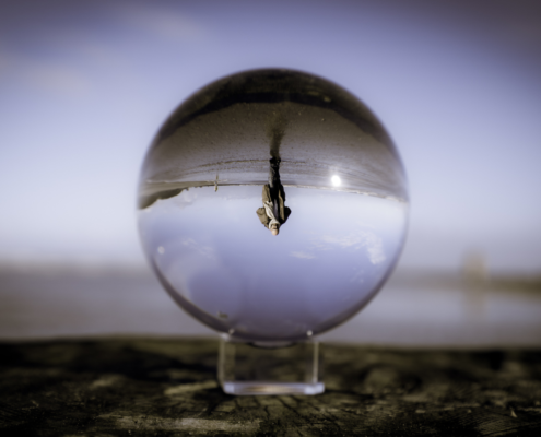 Das Bild zeigt einen Lensball, der alle Bilder auf den Kopf stellt. Er ist in der Mitte des Bildes und steht auf einem Holzpoller. Bis auf das Bild in der Kugel ist alles verschwommen. Dort sieht man Roland über den Strand gehen mit einem Blick nach vorne. Er steht dabei Kopf.