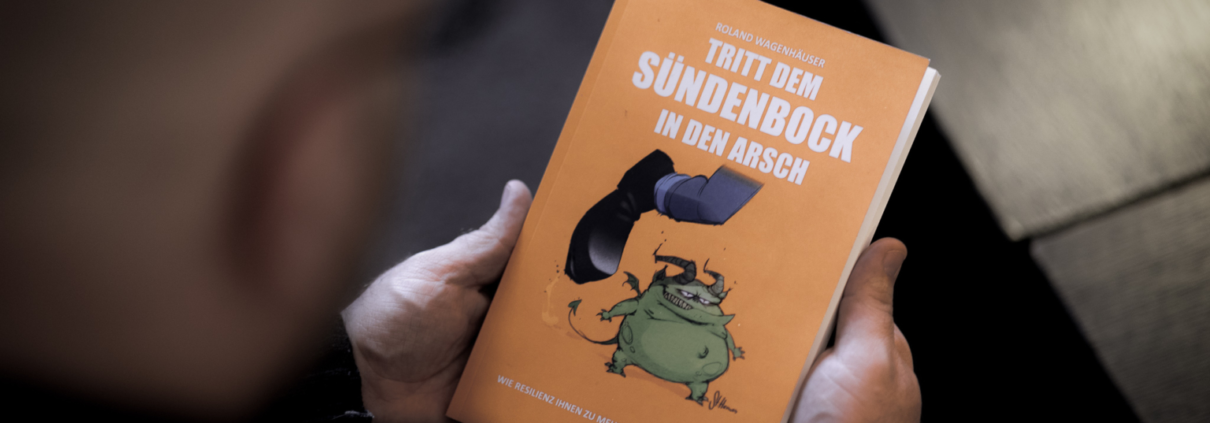 Im Fokus des Bildes ist das orange Buch "Tritt dem Sündenbock in den Arsch" von Roland Wagenhäuser. Der Blick geht direkt darauf und man sieht verschwommen den Hinterkopf von Roland und ein kleines Stück vom Tisch.