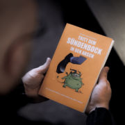 Im Fokus des Bildes ist das orange Buch "Tritt dem Sündenbock in den Arsch" von Roland Wagenhäuser. Der Blick geht direkt darauf und man sieht verschwommen den Hinterkopf von Roland und ein kleines Stück vom Tisch.
