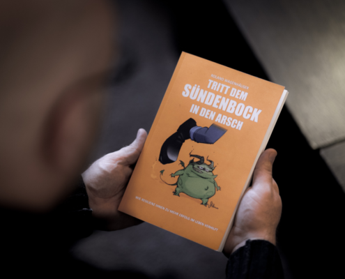 Im Fokus des Bildes ist das orange Buch "Tritt dem Sündenbock in den Arsch" von Roland Wagenhäuser. Der Blick geht direkt darauf und man sieht verschwommen den Hinterkopf von Roland und ein kleines Stück vom Tisch.