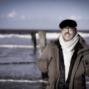 Das Bild zeigt Roland Wagenhäuser am Meer. Im Hintergrund sieht man Wellen an den Starnd kommen, und zwei Pfeiler im Meer. Roland Trägt eine schwarze Baskenmütze, eine Brille und einen dicken weißen Schal. Seine Hände hat er in die geöffnete Jacke gesteckt. Sein Blick geht nach vorne.