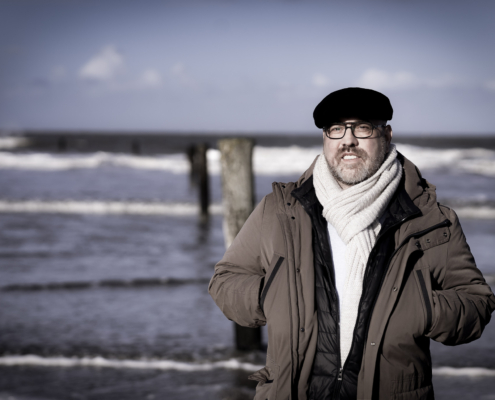 Das Bild zeigt Roland Wagenhäuser am Meer. Im Hintergrund sieht man Wellen an den Starnd kommen, und zwei Pfeiler im Meer. Roland Trägt eine schwarze Baskenmütze, eine Brille und einen dicken weißen Schal. Seine Hände hat er in die geöffnete Jacke gesteckt. Sein Blick geht nach vorne.